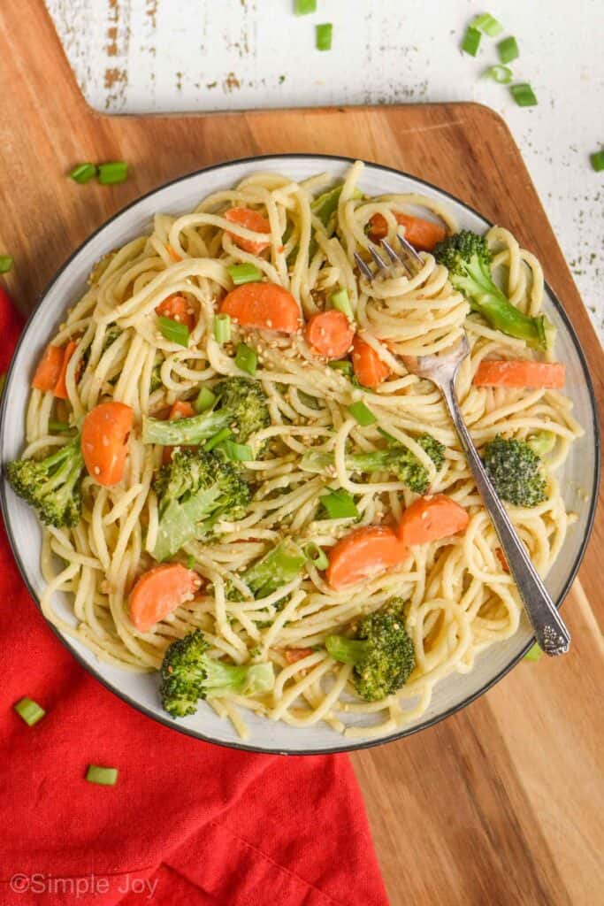 a plate filled with spaghetti mixed with carrots and broccoli, with a fork twirled with spaghetti on a cutting board with sliced green onion pieces