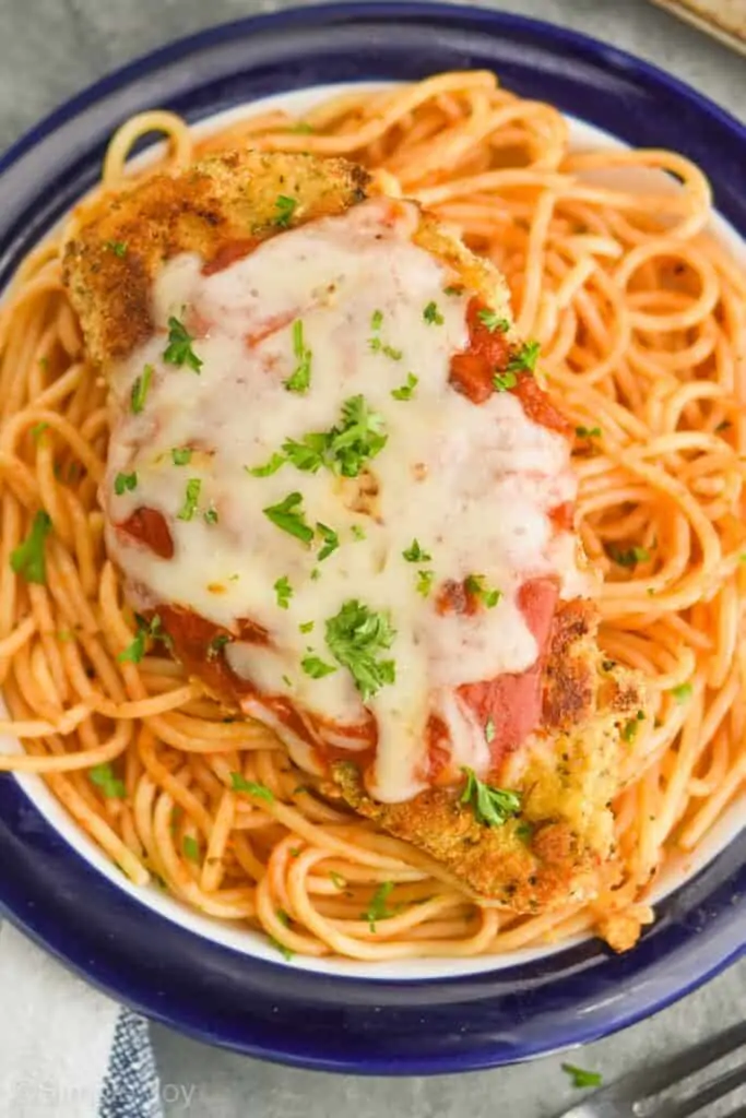close up overhead view of a piece of baked parmesan chicken on a bed of spaghetti garnished with fresh parsley