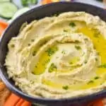 a black bowl full of hummus garnished with oil and parsley on a tray with vegetables, can see cut carrots, sliced cucumbers, and cherry tomatoes