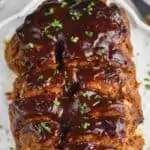 close up overhead view of a cooked meatloaf on a serving plate that has been slices, topped with more BBQ sauce, and garnished with parsley