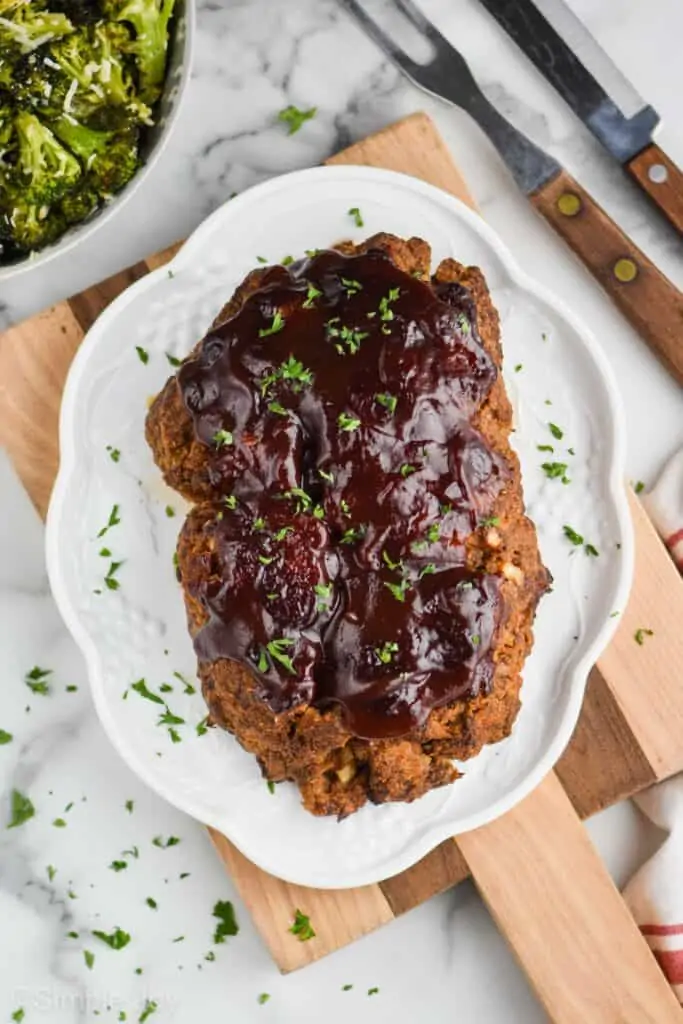 overhead view of an oval platter with cooked meatloaf on it topped with BBQ sauce and fresh parsley and a bowl of parmesan broccoli next it it