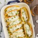 overhead view of a ceramic baking dish filled with 10 baked tacos that have white cheese melted on top, two beer mugs next to the casserole dish