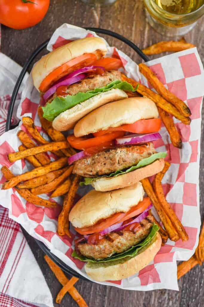 three grilled turkey burgers with lettuce, tomatoes, onions, and ketchup on their sides in a basket, surrounded by sweet potato fries