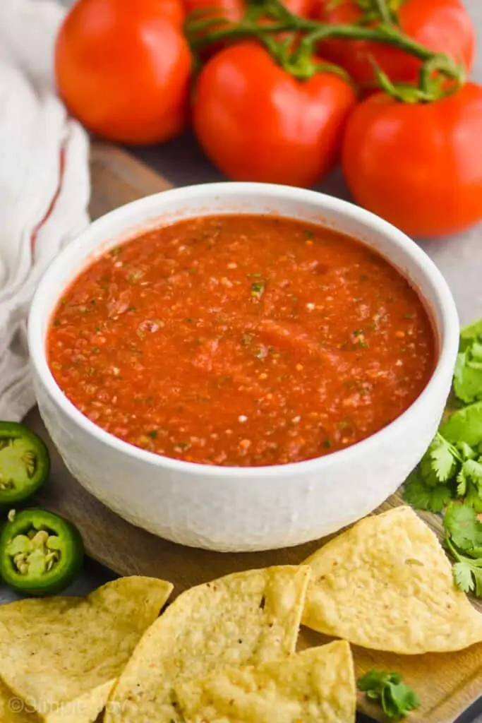 white bowl full of blender salsa, thin in texture, with tortilla chips in the foreground and tomatoes on the stem in the background