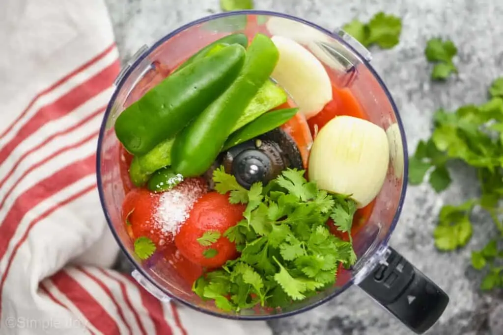 overhead view of a blender with unblended salsa ingredients: whole cilantro, a white onion cut in half, strips of jalapeño pepper sides, salt, and whole canned tomatoes
