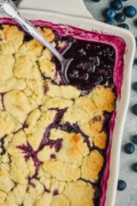close up overhead overhead a serving spoon in a ceramic dish filled with blueberry cobbler