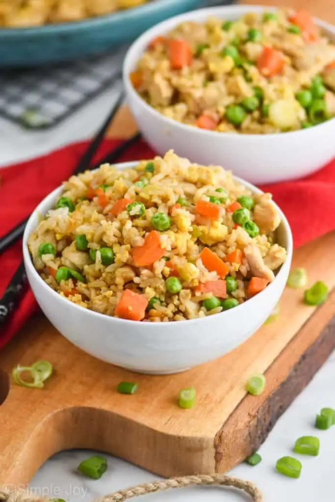 pulled back view of a white bowl filled with easy chicken fried rice recipe with peas, carrots, and eggs on a wooden cutting board with another bowl in the background