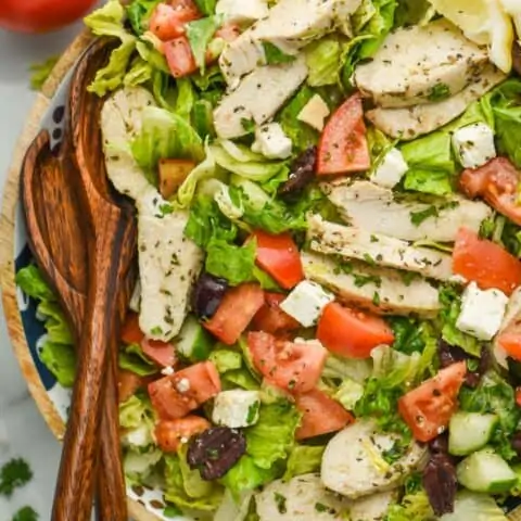 overhead view of a salad with tomatoes, squares of feta cheese, slices of chicken covered in herbs, kalamata olives, and two lemon wedges for garnish