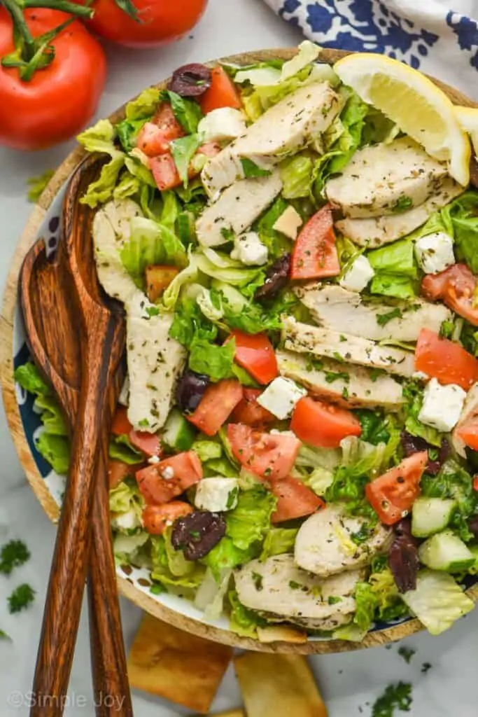 overhead view of a salad with tomatoes, squares of feta cheese, slices of chicken covered in herbs, kalamata olives, and two lemon wedges for garnish