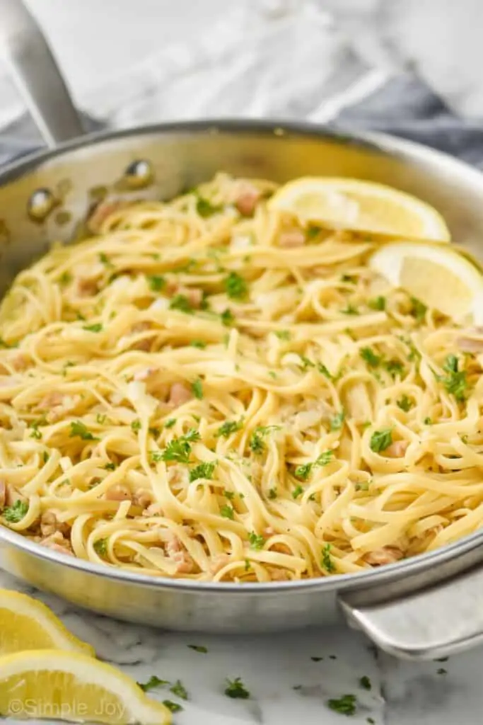 side view close up look at a large metal skillet filled with linguine with clam sauce recipe garnished with fresh parsley and two lemon wedges