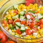 close up of corn salad in a glass bowl with fresh cut cucumbers, tomatoes, red onions, feta cheese, and fresh basil on top