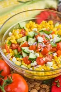 close up of corn salad in a glass bowl with fresh cut cucumbers, tomatoes, red onions, feta cheese, and fresh basil on top
