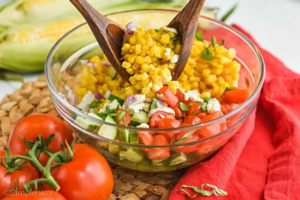 two wooden spoons tossing a glass bowl full of corn tomato salad