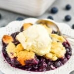 close up of a white dessert plate with blueberry cobbler recipe topped with vanilla ice cream, served with a spoon