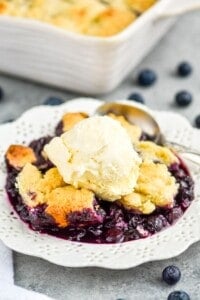 close up of a white dessert plate with blueberry cobbler recipe topped with vanilla ice cream, served with a spoon