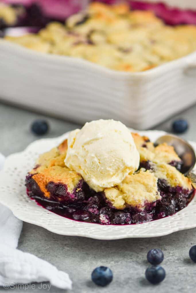 close up of a white dessert plate with blueberry cobbler recipe topped with vanilla ice cream, served with a spoon