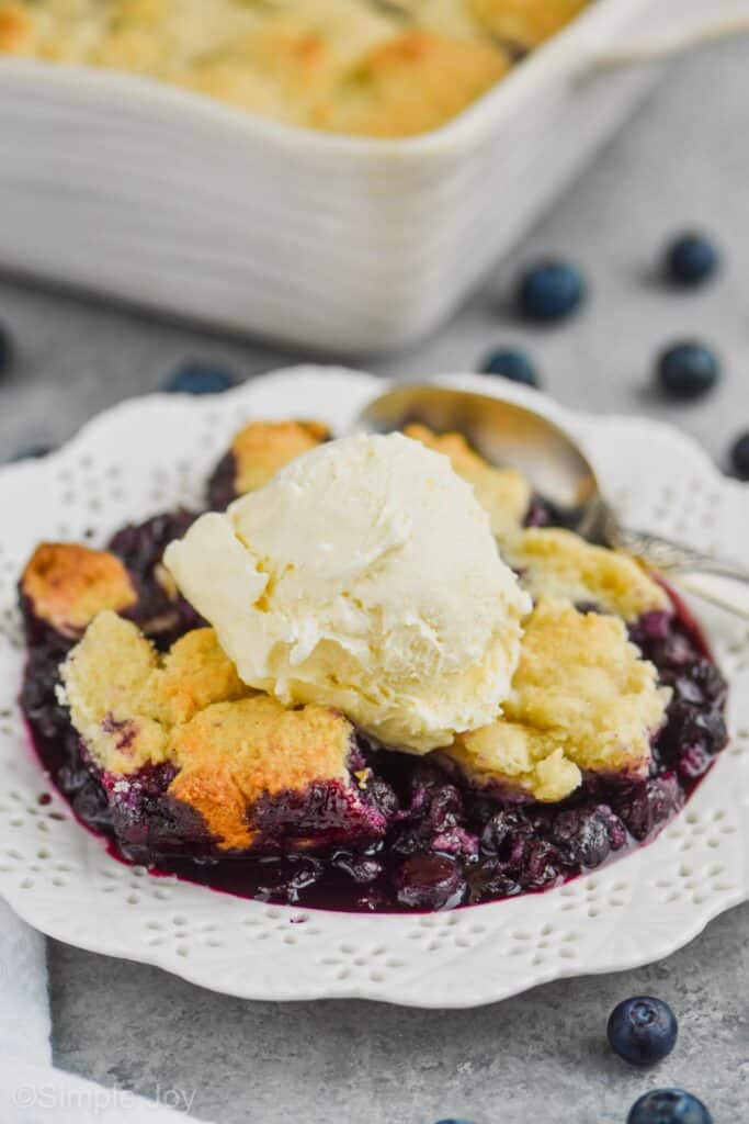 close up of a white dessert plate with blueberry cobbler recipe topped with vanilla ice cream, served with a spoon