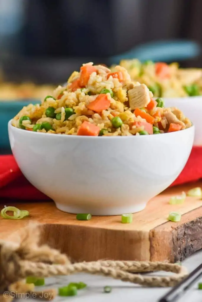 straight forward view of a white bowl full of a mound of chicken friend recipe recipe on a wooden cutting board against a dark black background with green onion slices around the cutting board