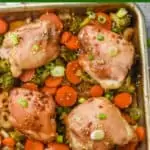 teriyaki chicken thighs on a baking sheet with broccoli, onions, and carrots, garnished with sesame seeds and green onions