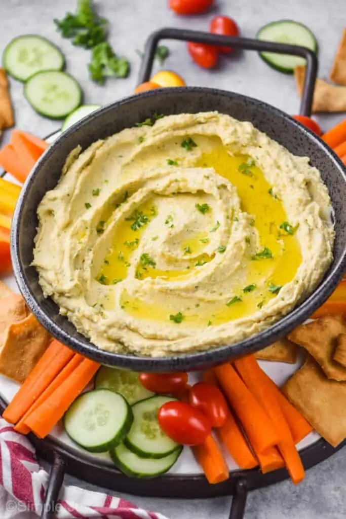 pulled back look of an odd shaped/oval black ceramic bowl full of easy hummus recipe garnished with olive oil and fresh parsley on a tray with handles on a tray with sliced fresh vegetables