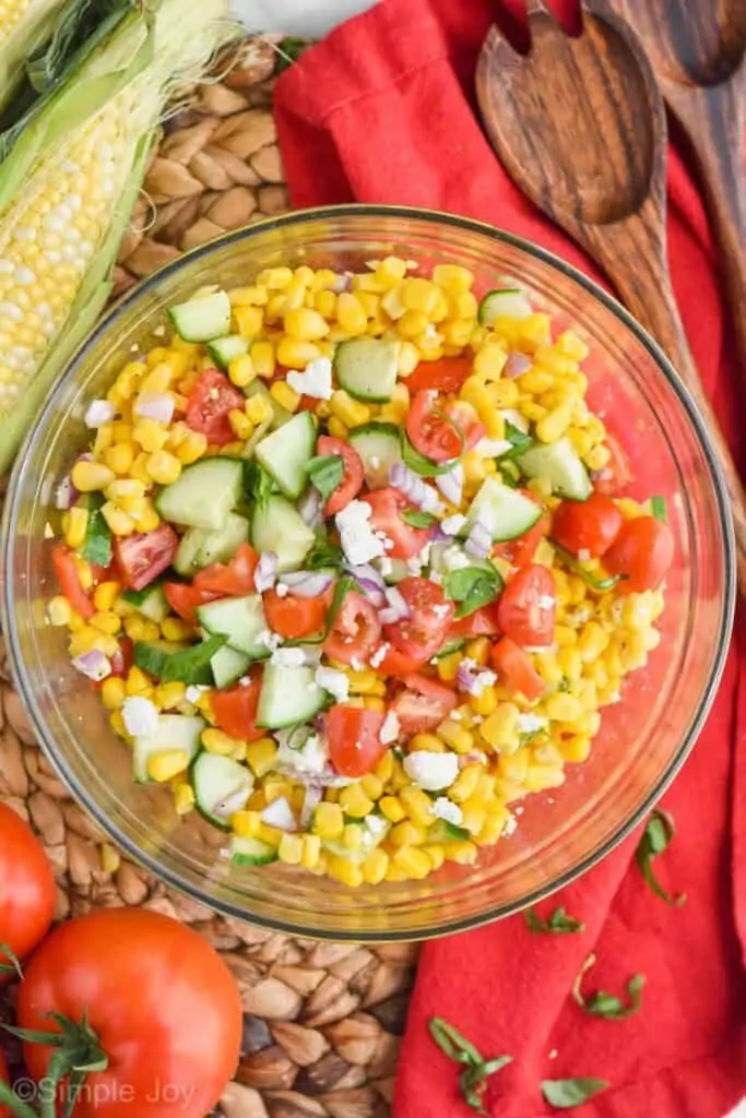 overhead of fresh corn salad in a glass bowl with fresh cut cucumber, tomato, red onion, feta and basil on top