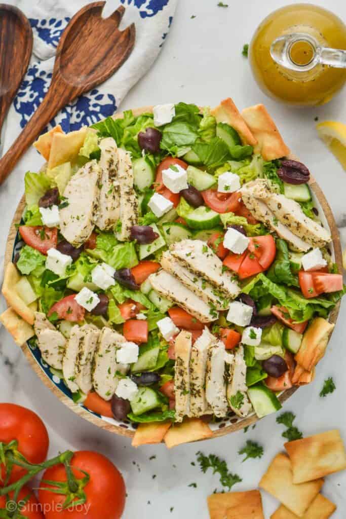 overhead view of a greek salad with chicken slices in a big serving bowl, garnished with fresh parsley, and pita chips off to the side