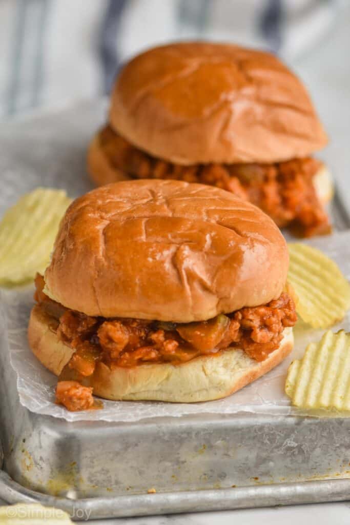 two healthy turkey sloppy joes sitting on wax paper on an overturned rimmed baking sheet with ruffled potato chips around it