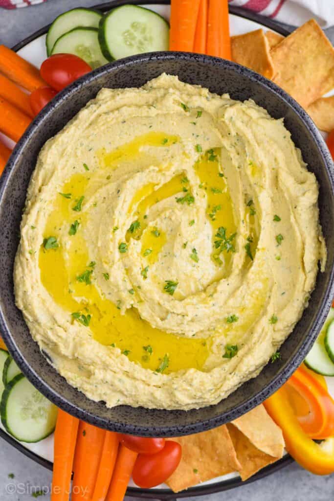 close up overhead view of an oval black bowl full of homemade hummus garnished with oil and fresh parsley on a tray with pita chips, carrot spears, pepper slices, slices of cucumbers, and cherry tomatoes