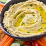 a black bowl full of hummus garnished with oil and parsley on a tray with vegetables, can see cut carrots, sliced cucumbers, and cherry tomatoes - 11