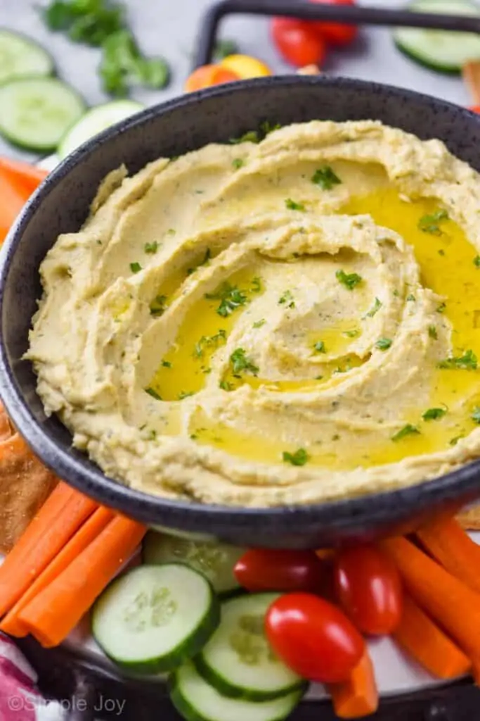 a black bowl full of hummus garnished with oil and parsley on a tray with vegetables, can see cut carrots, sliced cucumbers, and cherry tomatoes