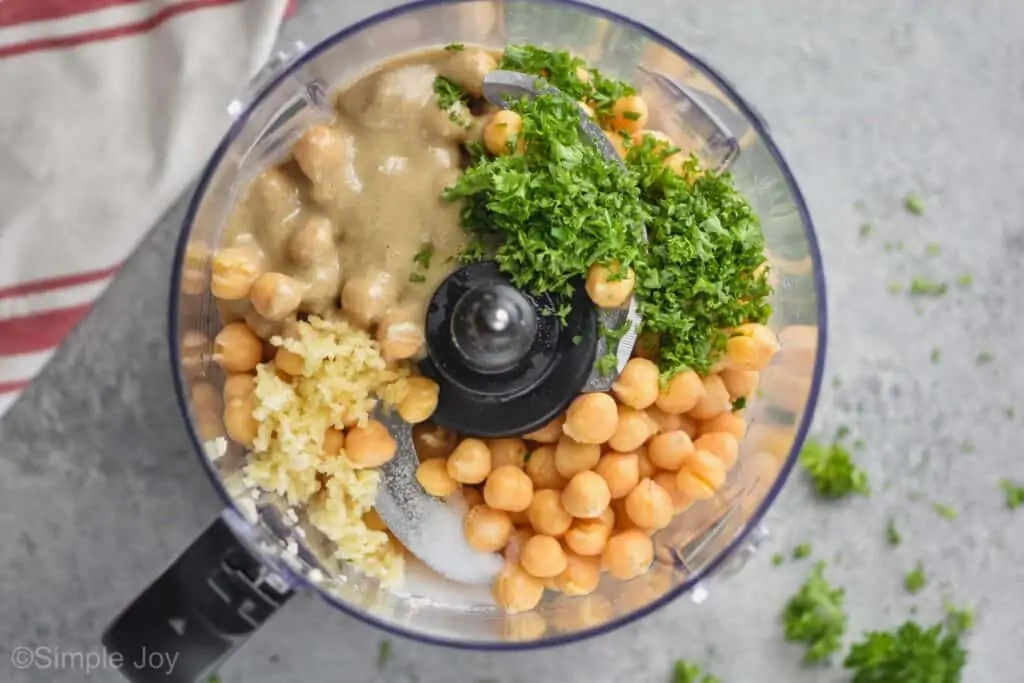 overhead view of a food processor with chickpeas fresh parsley minced garlic and tahini on a gray surface with spilled parsley and a cloth red and white napkin