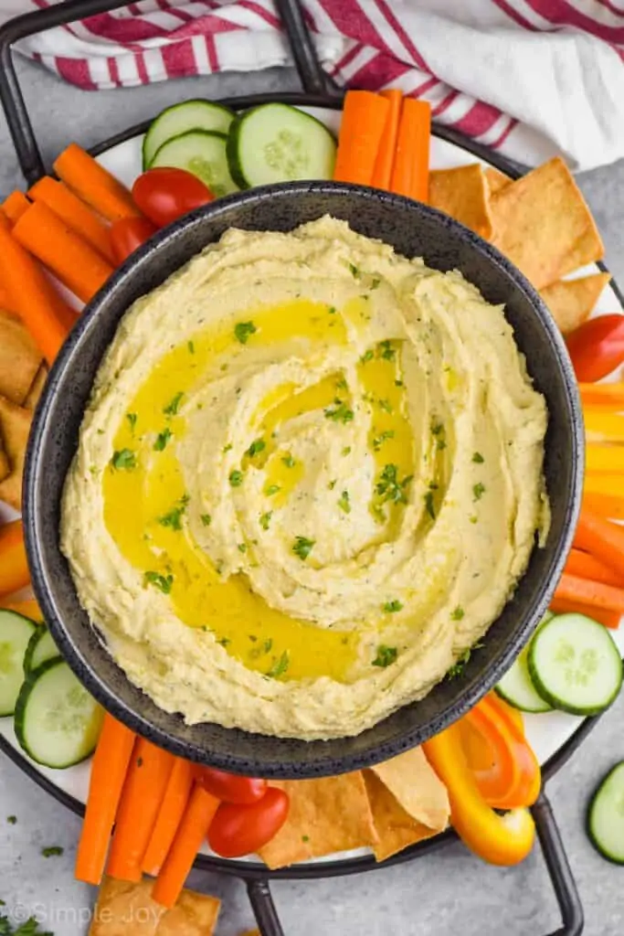overhead pulled back look at a black oval bowl full of hummus recipe garnished with olive oil and fresh parsley on a tray with cherry tomatoes, carrot pieces, slices of cucumbers, pepper slices, and pita chips on a gray surface with a white and red striped napkin