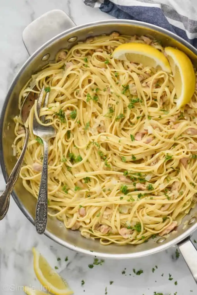 close up overhead view of a skillet with linguine and clams with a spoon and fork in in the pan, garnished with fresh parsley and two lemon wedges