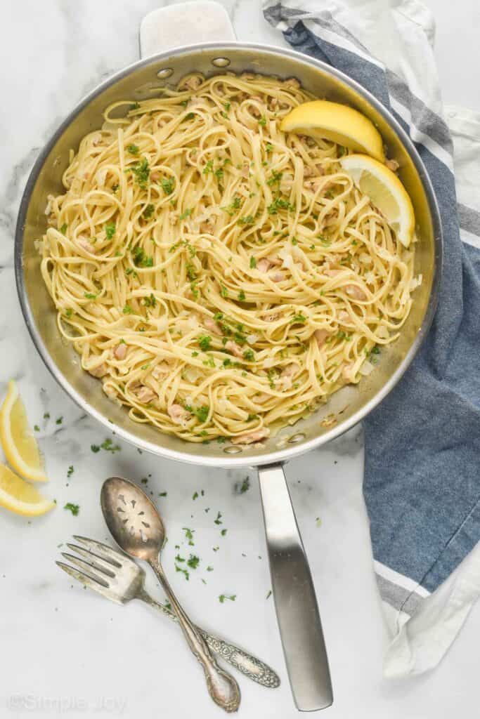 pulled back overhead view of a skillet filled with linguine with clam sauce, garnished with fresh parsley and two lemon wedges