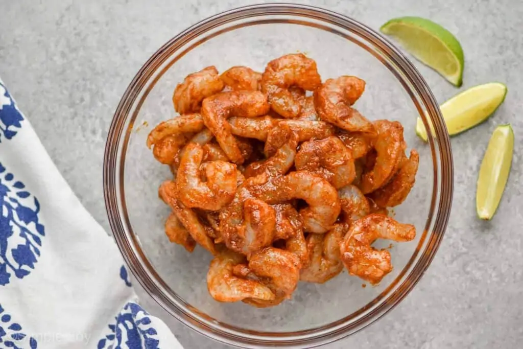 overhead view of a bowl of raw shrimp marinating in a combination of taco seasoning, oil, and lime juice with lime wedges next to the bowl