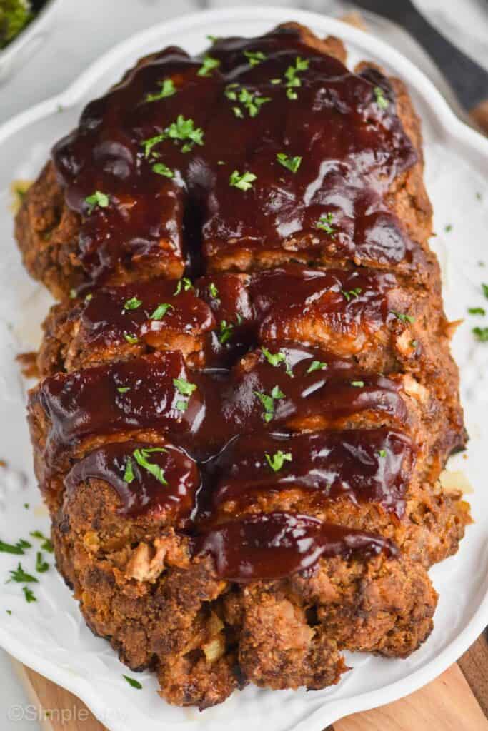 close up overhead view of a cooked meatloaf on a serving plate that has been slices, topped with more BBQ sauce, and garnished with parsley