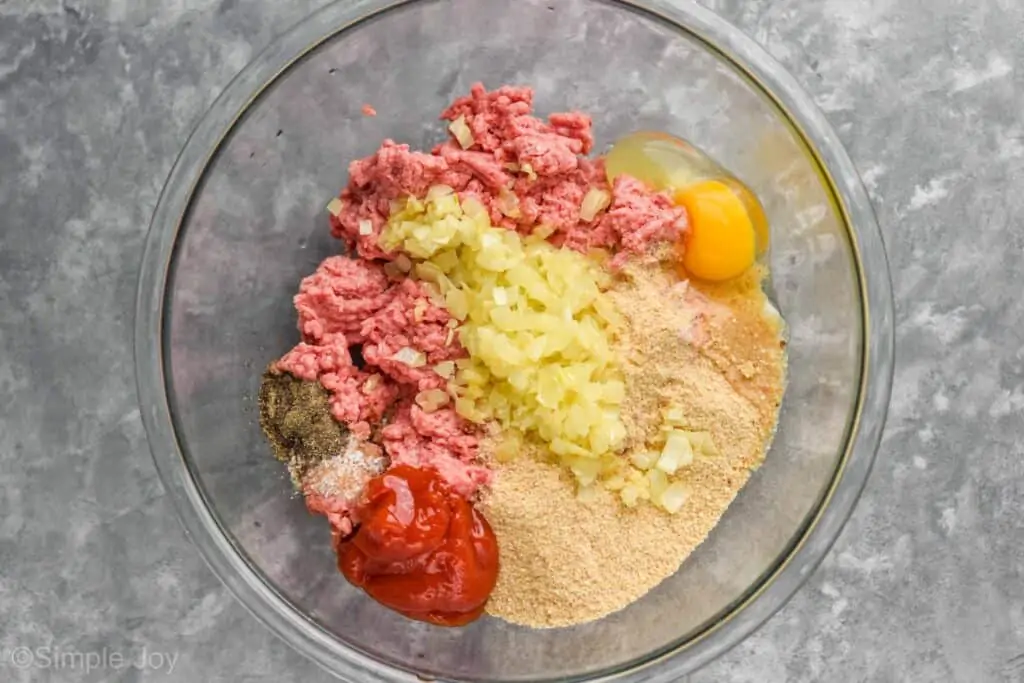 overhead view of a the ingredients for meatloaf: ground beef, raw egg, breadcrumbs, catchup, seasonings, and cooked onion