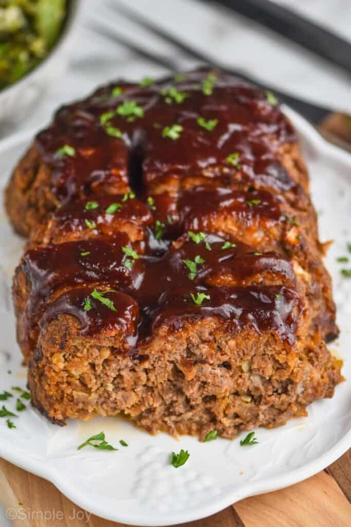 picture of a meatloaf on a serving plate, cut into with BBQ sauce on top and garnished with fresh parlsey