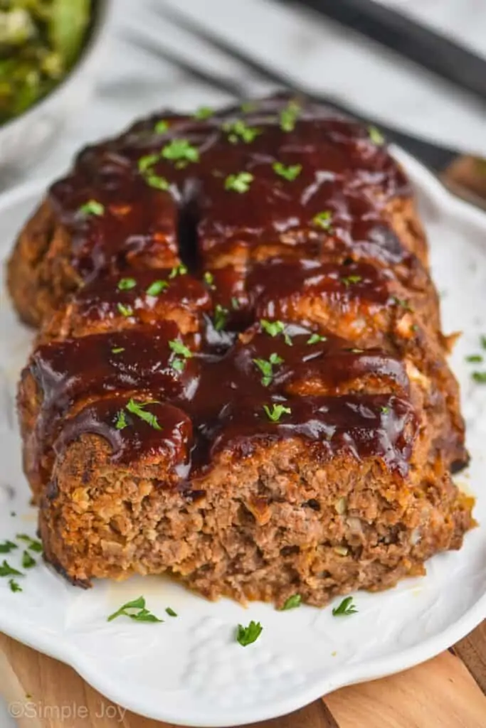 picture of a meatloaf on a serving plate, cut into with BBQ sauce on top and garnished with fresh parlsey