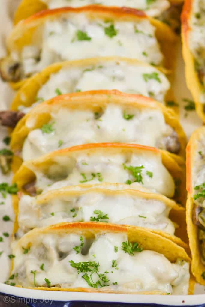 close up of baked tacos in a baking dish with melty white cheese, in hardshell tacos, and garnished with parsley