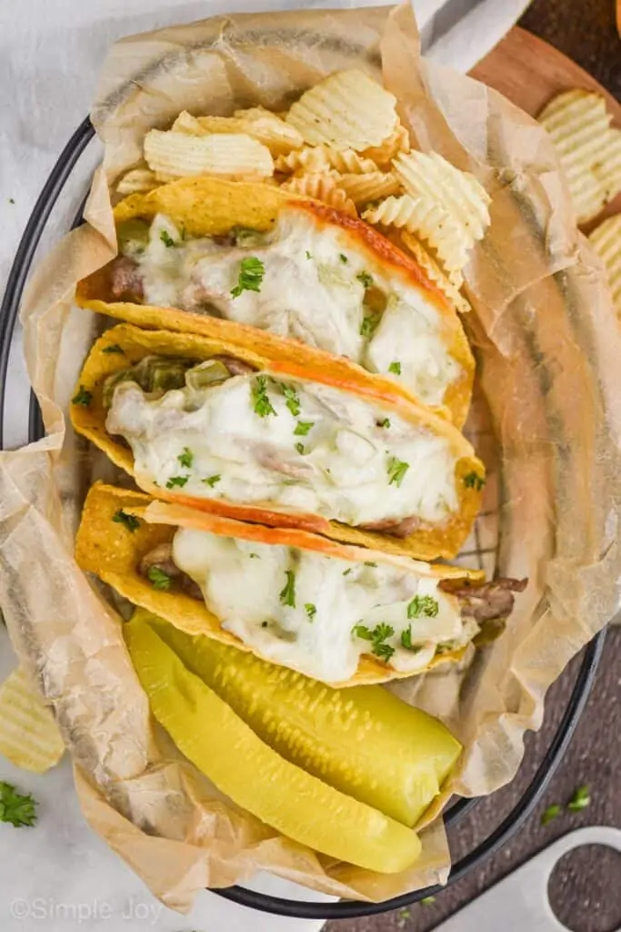 overhead view of a wire basket with parchment paper, three Philly cheesesteak baked tacos garnished with fresh parsley, two pickles and chips in the basket