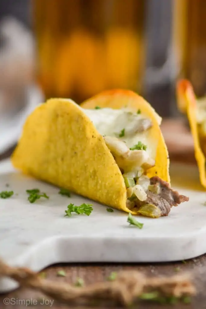 a Philly cheesesteak baked taco on a marble cutting board with a piece of steak coming out, provolone melted on top, and fresh parsley on top