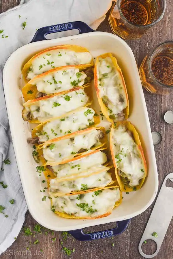 overhead view of a ceramic baking dish filled with 10 baked tacos that have white cheese melted on top, two beer mugs next to the casserole dish