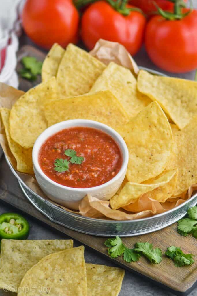 a small white bowl of salsa recipe garnished with cilantro in a metal tin with tortilla chips
