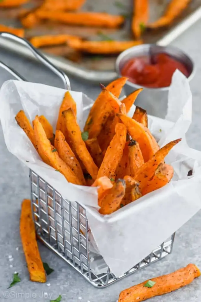 sweet potato fries in a wire basket that is lined with wax paper with a small cup of ketchup in the background
