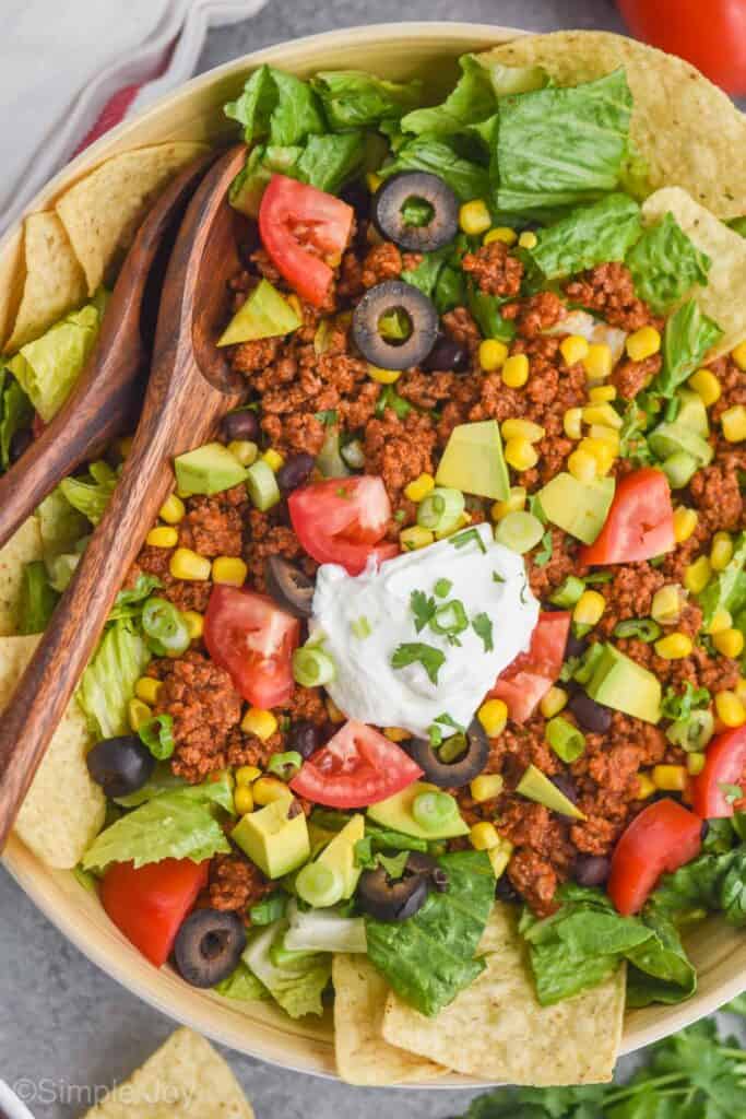 close up overhead view of taco salad with meat, corn, avocado, tomato, and a dollop of sour cream with wooden spoons for serving
