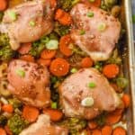 teriyaki chicken thighs on a baking sheet with broccoli, onions, and carrots, garnished with sesame seeds and green onions