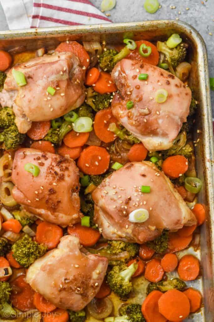 teriyaki chicken thighs on a baking sheet with broccoli, onions, and carrots, garnished with sesame seeds and green onions