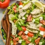 overhead view of a salad with tomatoes, squares of feta cheese, slices of chicken covered in herbs, kalamata olives, and two lemon wedges for garnish