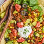 close up overhead view of taco salad with meat, corn, avocado, tomato, and a dollop of sour cream with wooden spoons for serving
